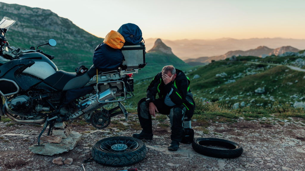 A concerned motorbike rider is sitting on the road in the middle of nowhere with his head in hand. He tried to change the tire but he failed